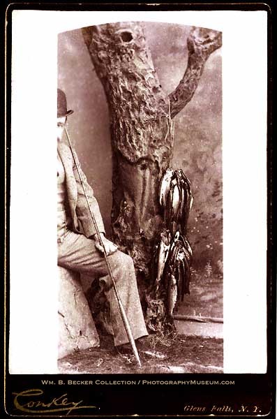 George W. Conkey, “A Day’s Catch,” c. 1890 — a seated angler with his face cropped from view, holding up a string of fish beside a studio tree